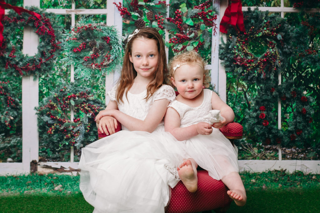 2 girls smiling for a Christmas portrait with a festive wreath wall background by Hobart Christmas Photo expert Lauren Vanier
