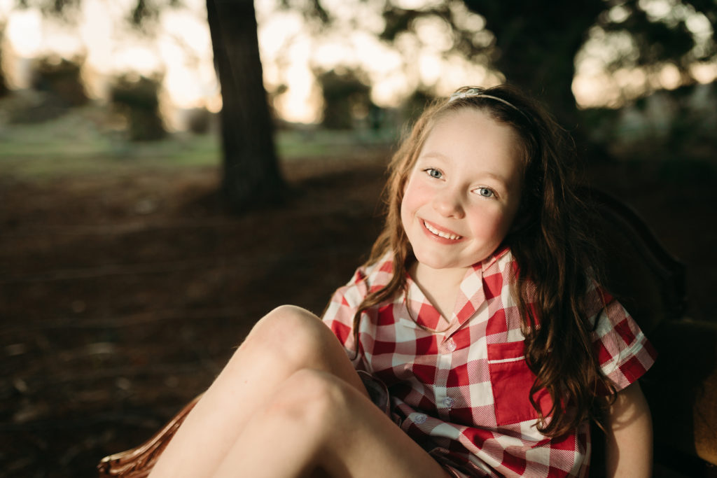 Girl in Christmas Pajamas smiling for the camera with the sunset behind her through the trees with bokeh. By Lauren Vanier Photography in Hobart, Tasmania