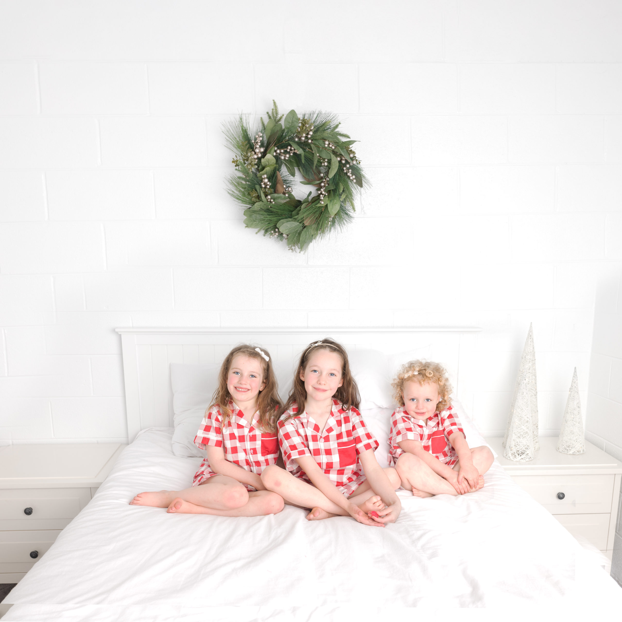 3 children sitting on a bed posing for a christmas themed photoshoot while wearing christmas pajamas. There is a wreath on the wall in the background and the photo is captured by Lauren Vanier Photography in Hobart