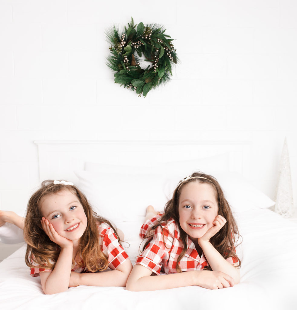 Christmas Mini Session image with 2 girls wearing Christmas pajamas on a while bed set up with a Christmas wreath on the wall in the background.  The girls are smiling and captured by Hobart Portrait photographer Lauren Vanier